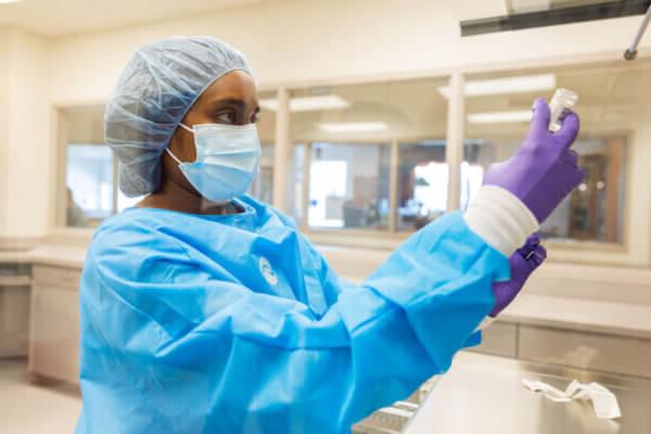 十大正规博彩网站评级博彩平台推荐大学 Bernard J. Dunn 药学院 student in blue scrubs, gloves, cap and mask, holding a vial.